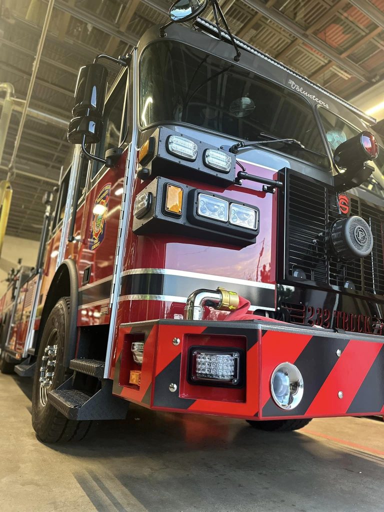 A red fire truck parked inside a fire station, featuring shiny reflective surfaces, multiple lights on the front, and black and yellow striping accents. The garage ceiling and parts of other fire station equipment are visible in the background.