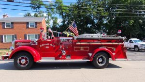 A vintage red fire truck from Pleasant Hill with "1951" marked on it is parked on a residential street. The truck has shiny chrome details, an American flag, and fire equipment on the sides. Trees and brick houses are in the background.