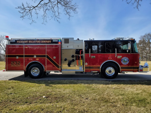 A red fire truck with "Pleasant Hills Fire Company" and "232 Engine" written on the side is parked on a grassy area next to a road. The side compartments are open, revealing equipment inside. The sky is clear, and there are trees without leaves in the background.