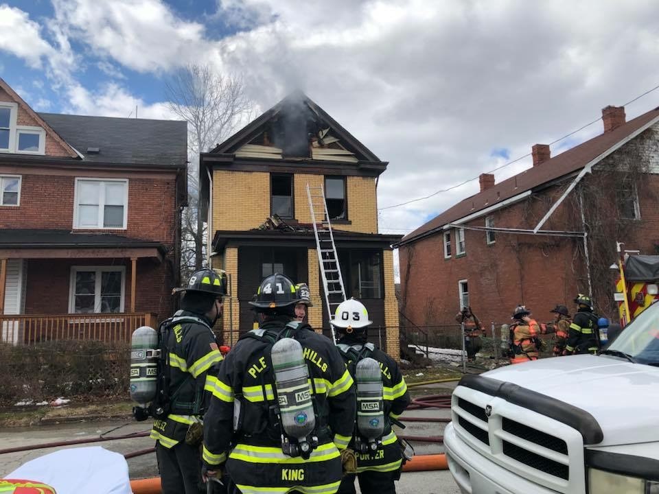 Firefighters in full gear work at the scene of a house fire in a residential area. Smoke rises from the top windows of a yellow brick house, and a ladder is positioned against the front. Nearby houses are visible, and other emergency personnel are also present.