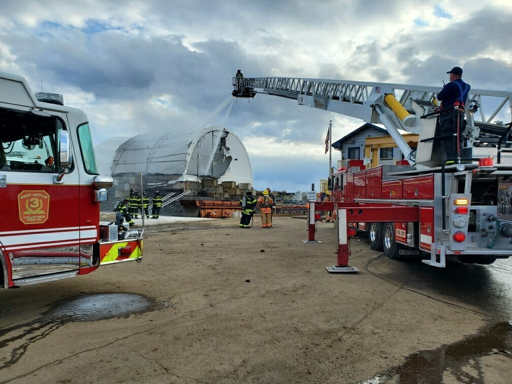 Firefighters in uniform work to extinguish a fire at a structure with a partially collapsed roof. Two fire trucks are present, one with a ladder extended and a firefighter spraying water from above. Another firefighter stands on the ground observing the scene.